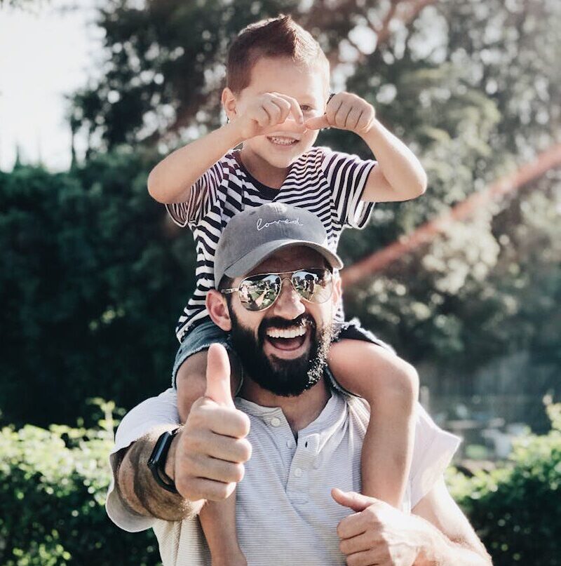 Father giving son a ride on shoulders, both laughing and smiling in a park setting.