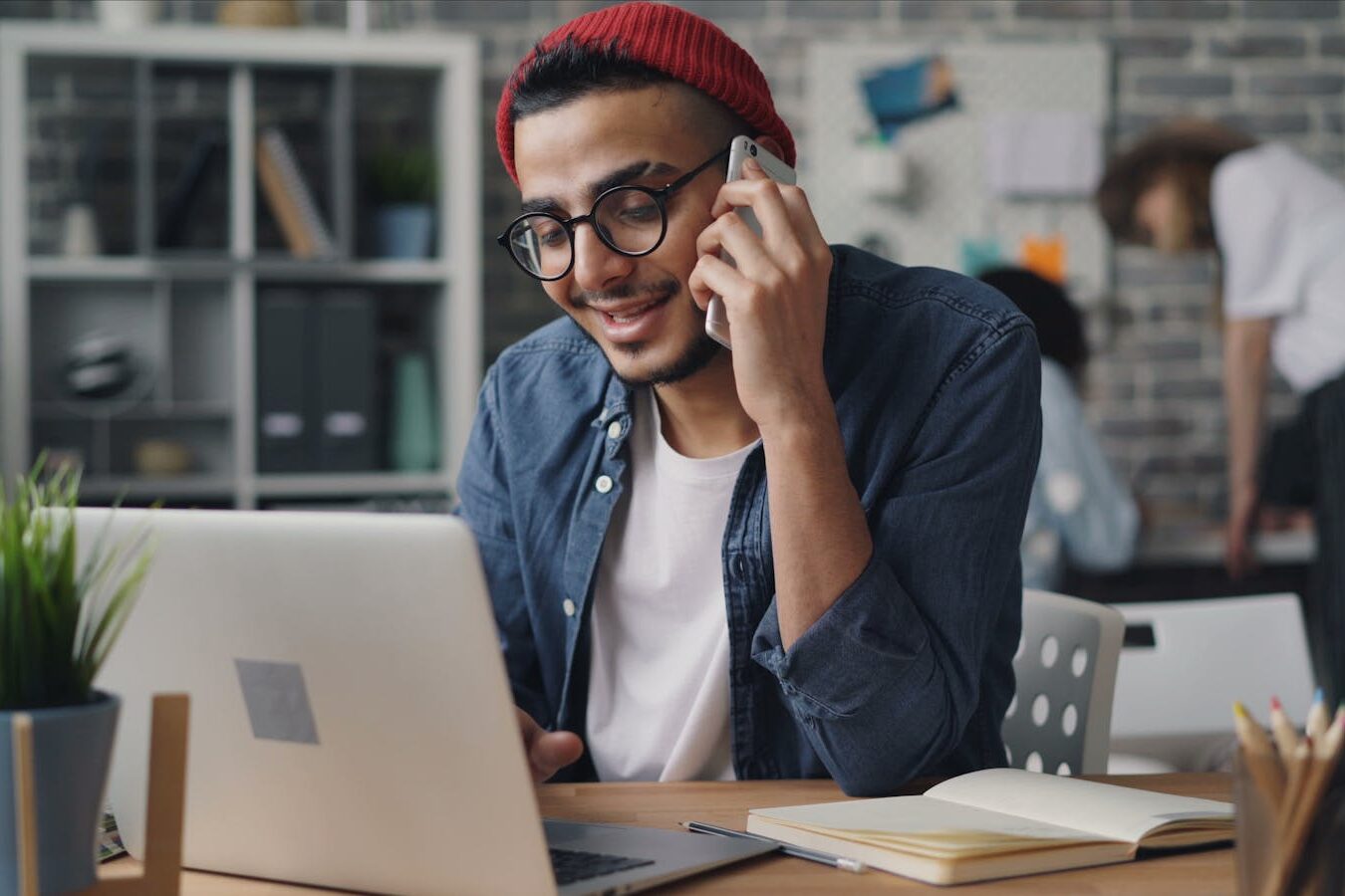 A young man in a modern office setting works on a laptop while talking on a smartphone.