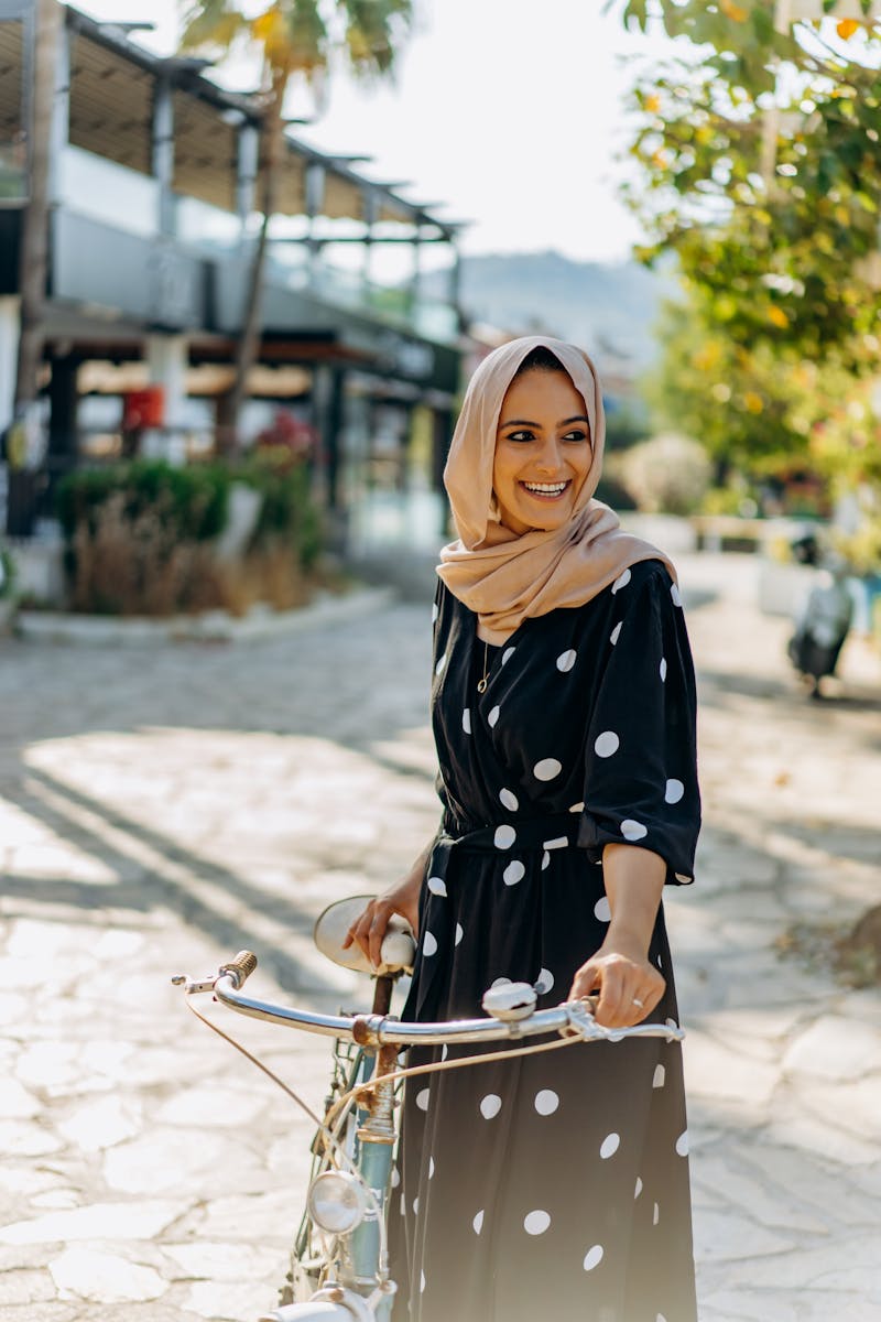 Young woman in polka dot dress and hijab smiling while holding a bicycle on a sunlit street.